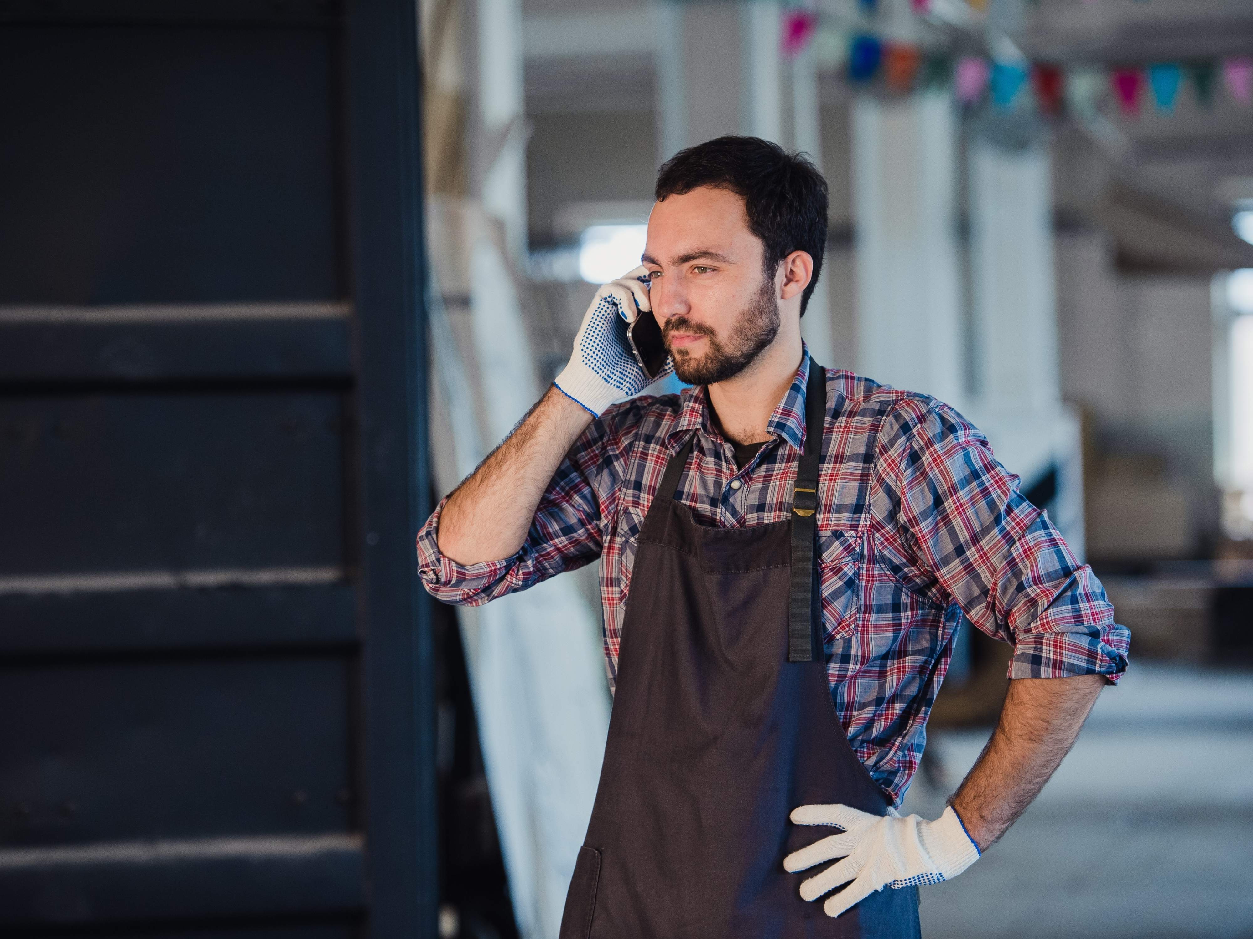 A bearded man in a flannel shirt and apron talks on the phone inside a workshop, possibly coordinating a job. His professional yet casual appearance reflects the personal touch often highlighted in garage door marketing strategies targeting local homeowners